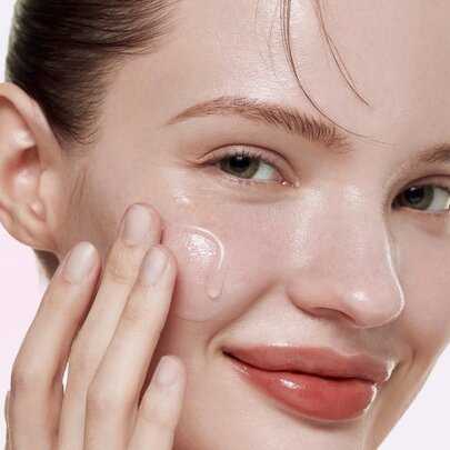 Model applying cream to her cheek, wearing shiny lipstick.