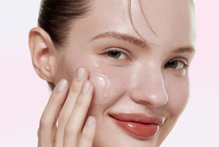 Model applying cream to her cheek, wearing shiny lipstick.
