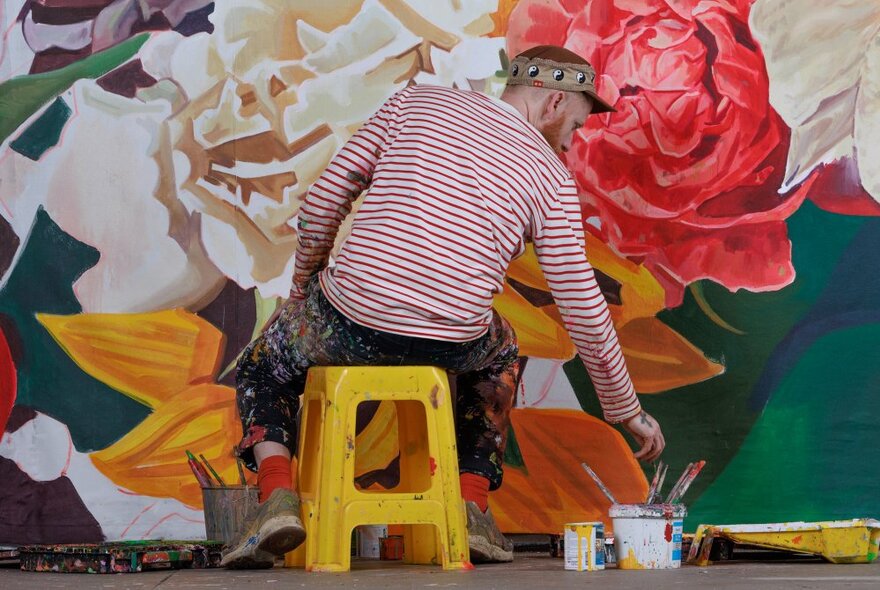 The back of an artist, sitting on a small yellow stool, painting a large mural of giant flowers on a wall. 