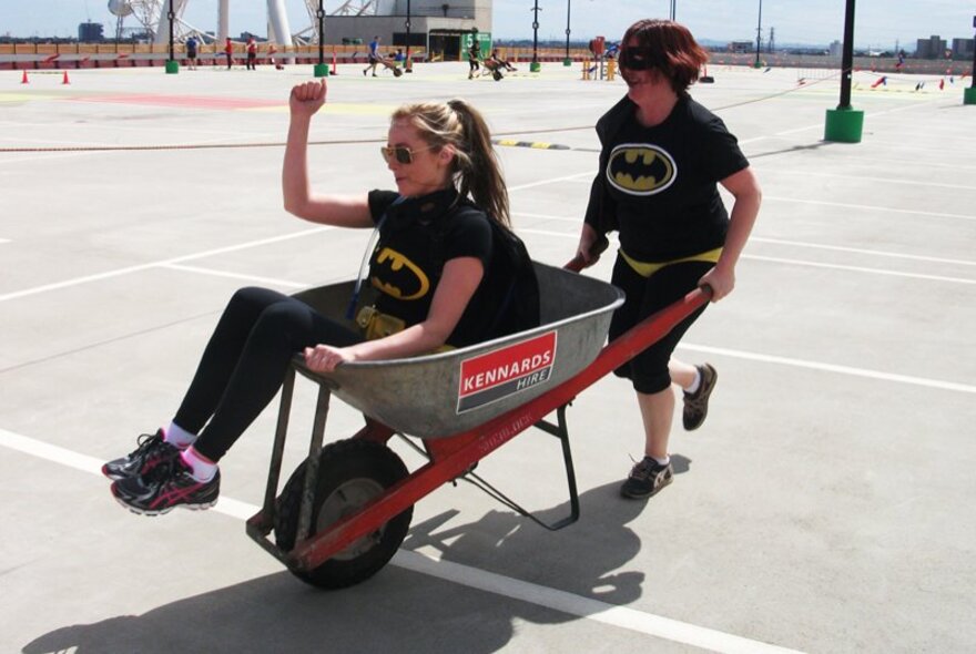 A person pushing another person in a wheelbarrow, across an empty parking lot in the city during the day, taking part in a challenge race.