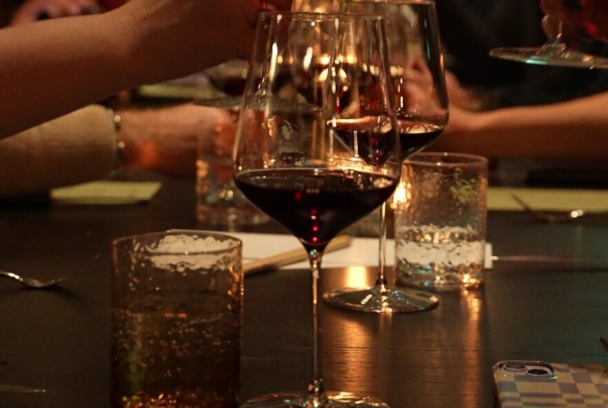 Glasses of wine and small candles on a restaurant dining table, with people's forearms visible in the background.
