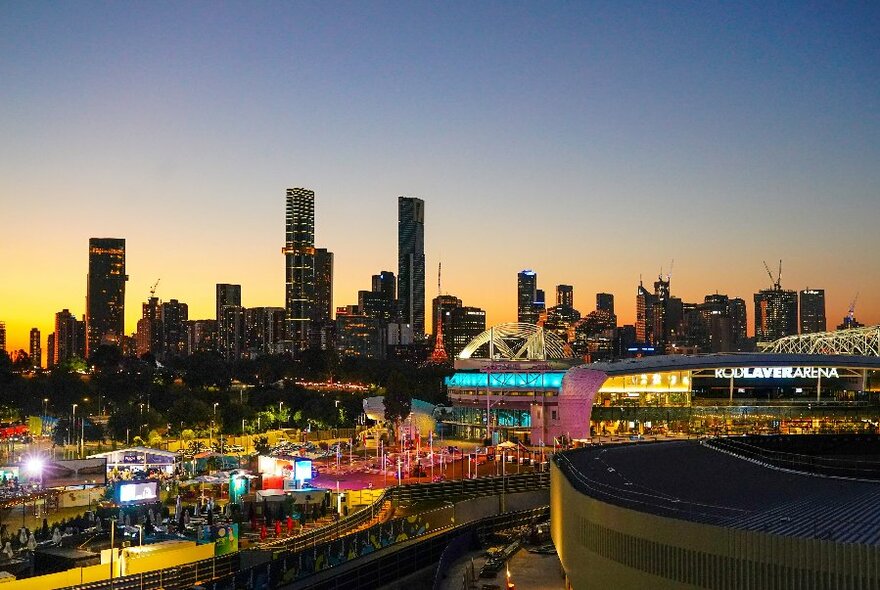 Melbourne Park arenas lit up at twilight, with city building skyline in the background.