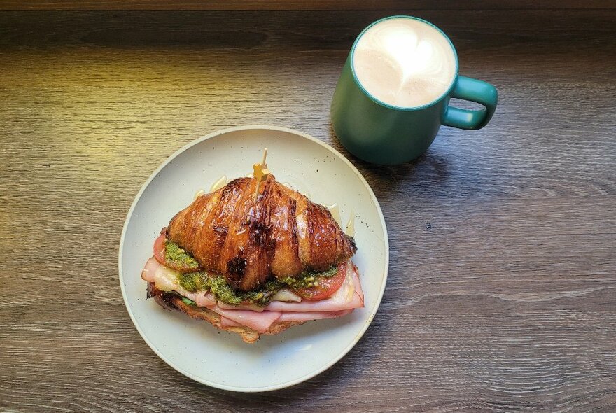 A ham, tomato and cheese croissant on a plate next to a mug of hot chocolate. 