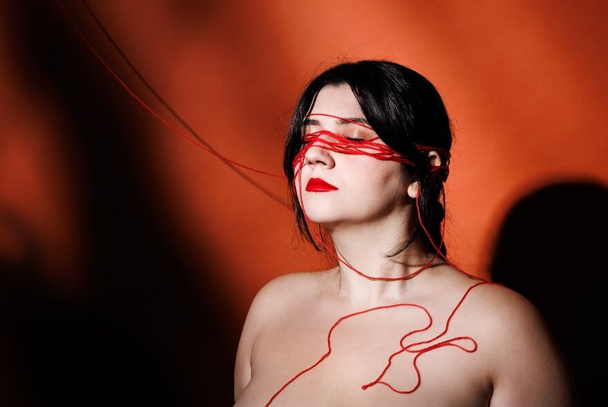 The naked upper torso of a woman with red string covering her closed eyes and curling over her shoulder.