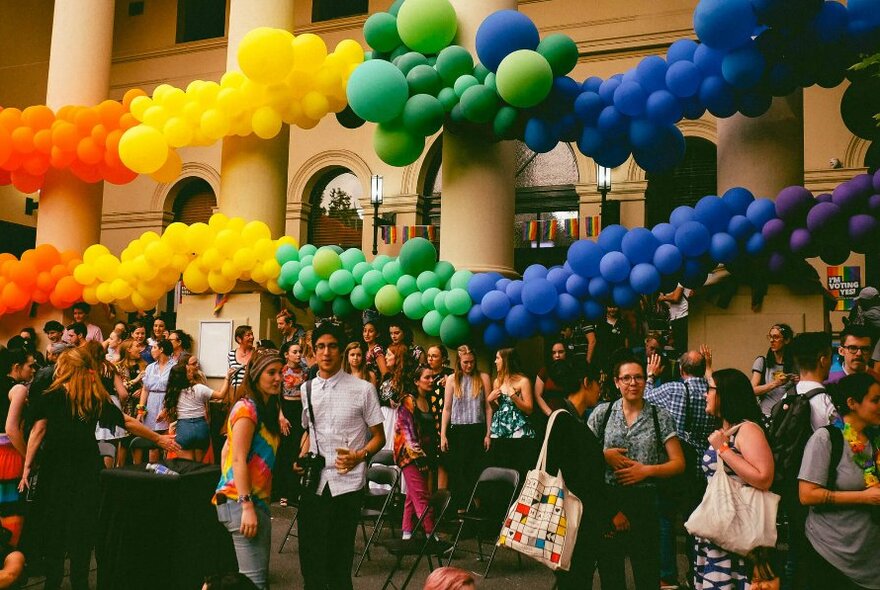 Balloons in coloured groups along a row of pillars, people milling below.