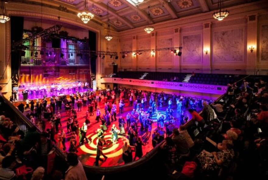 Looking down inside Melbourne Town Hall at many dancers on the floor which is lit in blue, red and pink. 