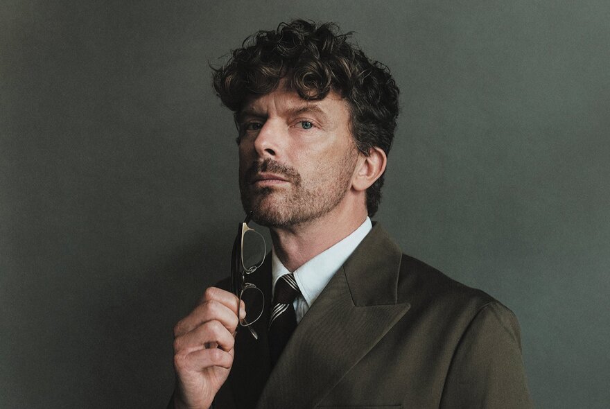 An actor in a suit, shirt and tie, holding a pair of glasses under his chair, with curly brown hair, looking serious.