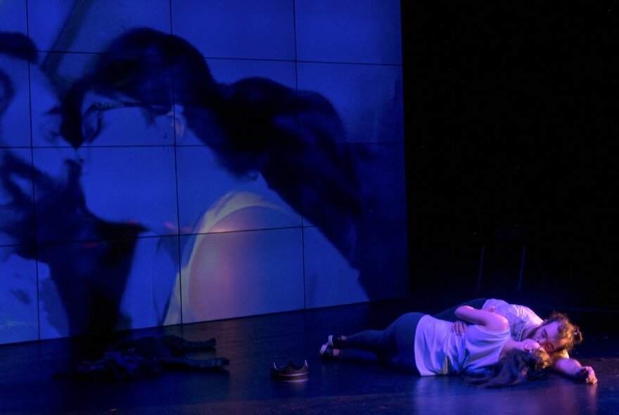 Two performers lying down on stage, with the image of them kissing as viewed from above projected onto a screen behind them.