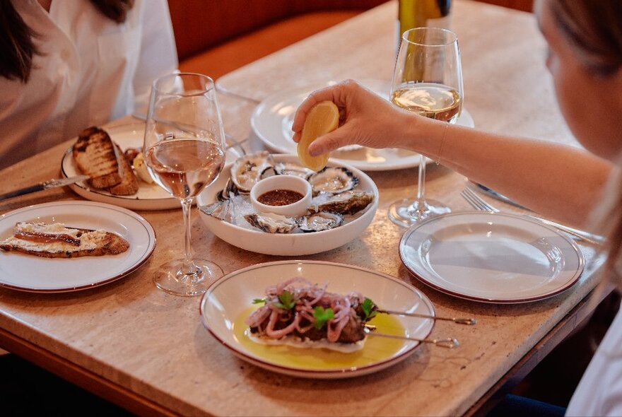 Two people sitting at a wooden table, one of them squeezing lemon over a plate of oysters, with other plates of food and two wine glasses.