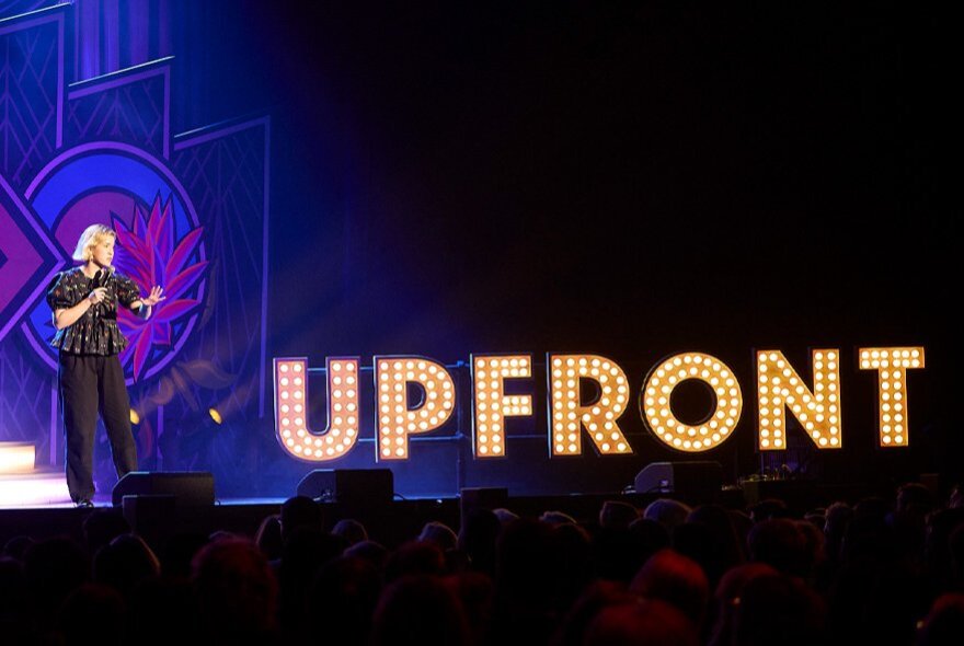 A comedian performing on a stage, holding a microphone, with a stage backdrop spelling out the words UPFRONT in bright lights.