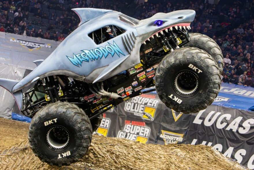 A monster truck with a shark face called Megaladon tips onto its back wheels in an arena.