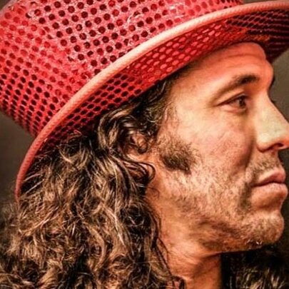 International magician Hernán Maccagno in side profile, wearing a red sequined top hat with his curly long brown hair visible.