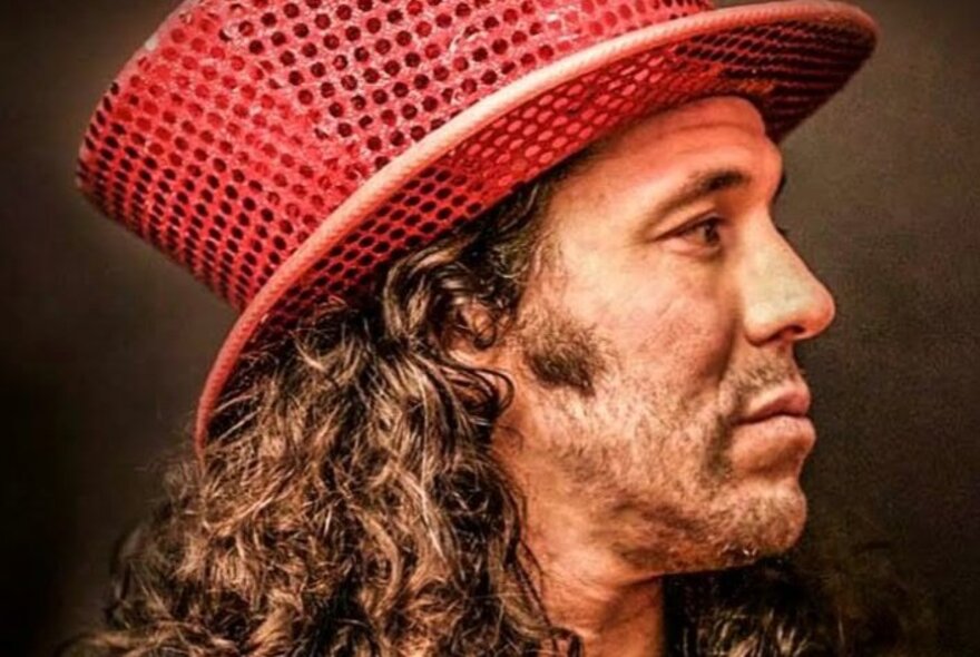 International magician Hernán Maccagno in side profile, wearing a red sequined top hat with his curly long brown hair visible.