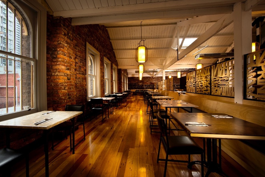 The interior of B'Churrasco, showing exposed brick wall, rows of tables and chairs, hanging pendant lights and a wood panelled ceiling.