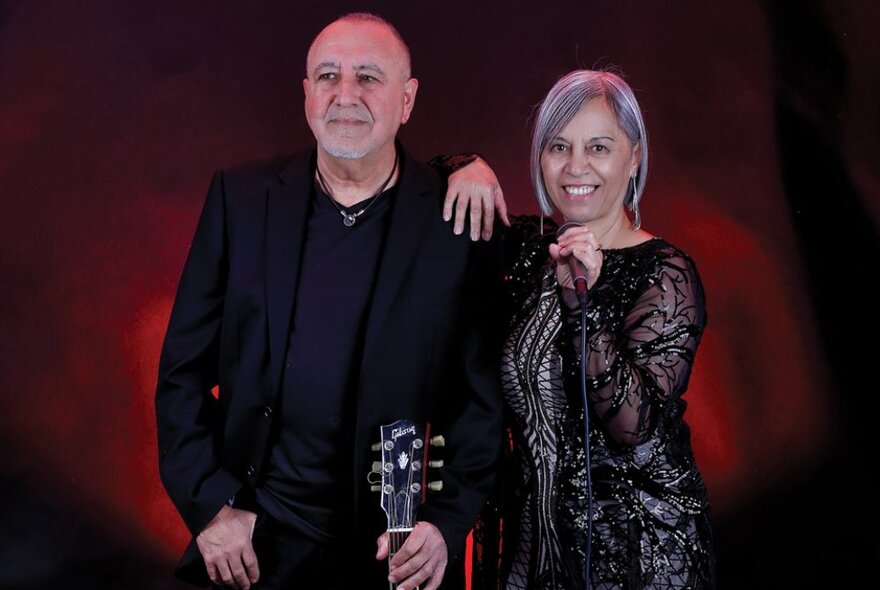 Male musician holding an acoustic guitar standing next to a female singer holding a microphone, her arm resting on his shoulder.