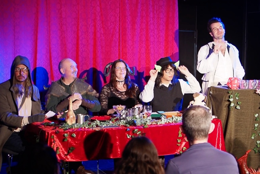 Performance team of five wearing costumes, on a small stage with red curtains, in front of a seated audience.