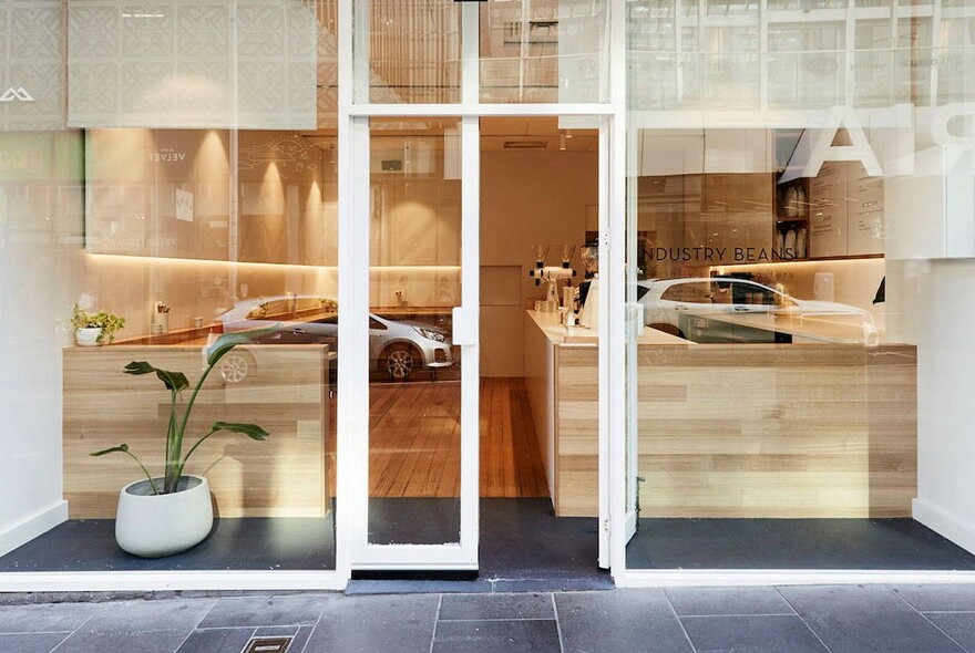 Shop exterior looking into the cafe's blonde-wood counters, with pot plant and grey tiled floors.