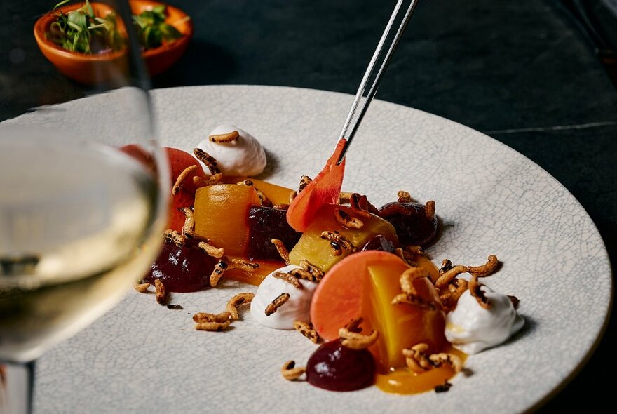 Tweasers adding sliced orange segments to a dish of food, with a glass of white wine in the foreground.