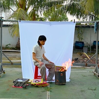 A person sitting in a backyard with a white backdrop hanging up behind them and a small fire in a tin beside them. 