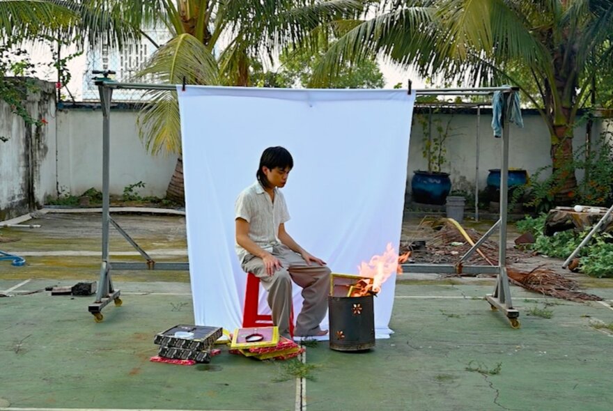 A person sitting in a backyard with a white backdrop hanging up behind them and a small fire in a tin beside them. 