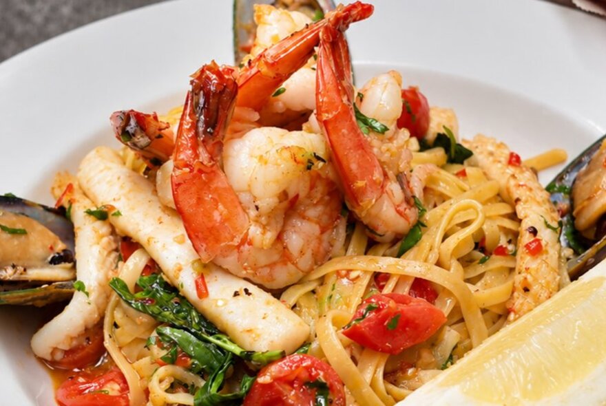 A seafood linguine pasta dish featuring large prawns, mussels, and diced tomatoes in a sauce, garnished with fresh herbs.