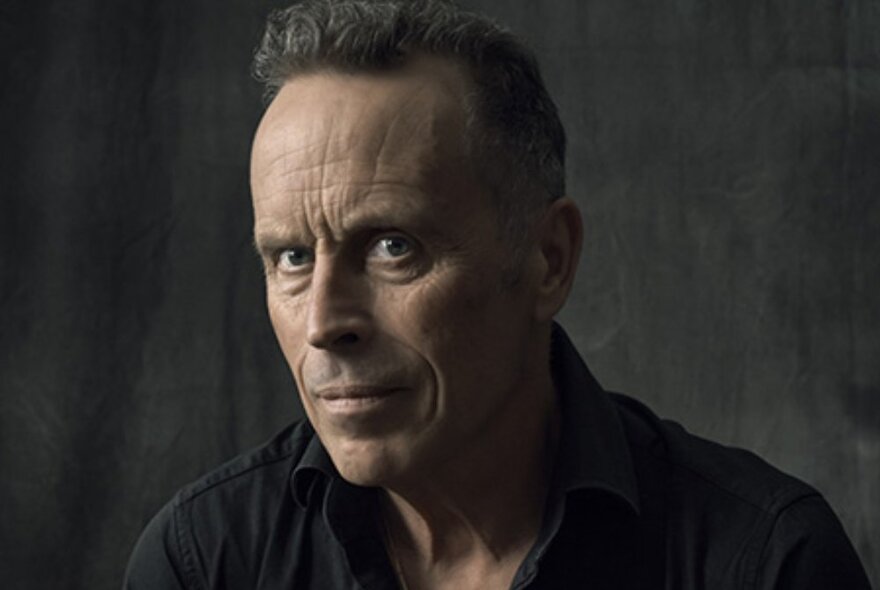 Musician Mark Seymour, frowning, wearing dark clothes against dark background.