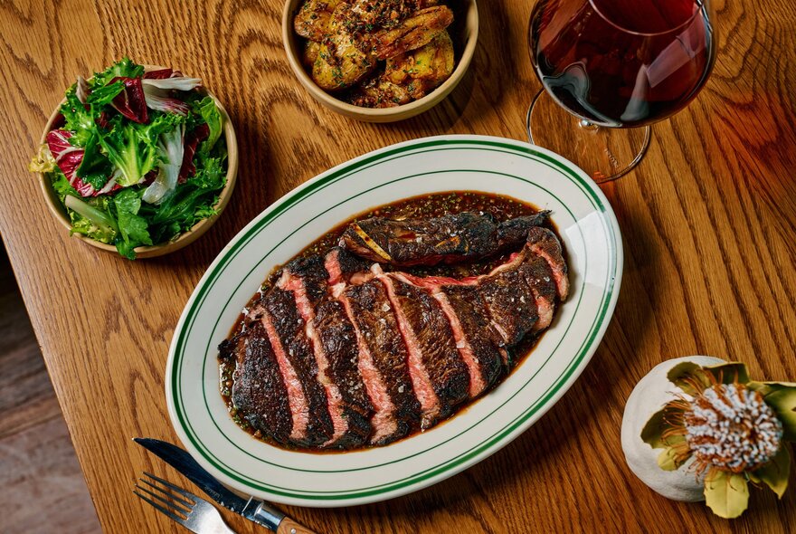 A plate of sliced rare steak alongside separate bowls of salad and potates on a wooden table, seen from above.