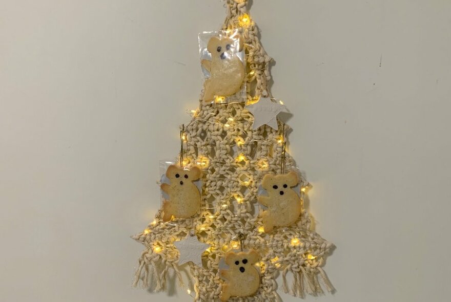 A flat macrame Christmas tree with small koala shaped decorations and fairy lights pinned to it, hanging on a wall.