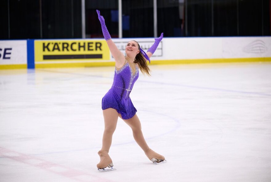 A female figure skater in a purple costume executing a dance move on the ice.