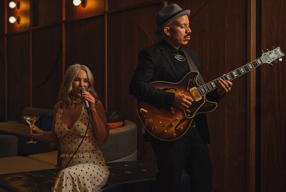 A man wearing a suit and hat stands playing guitar while a woman seated nearby sings into a microphone while holding a martini in her other hand.