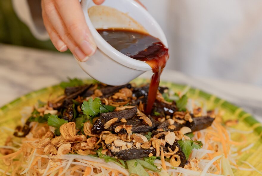 A hand pouring a dark sauce from a white ceramic bowl over a Vietnamese salad.