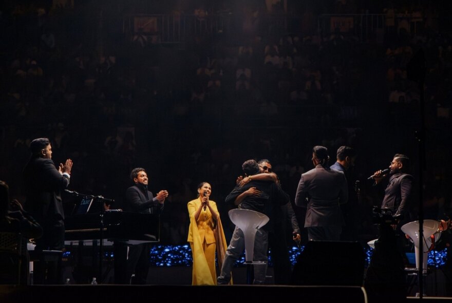 A group of performers and musicians on a dimly lit stage, with a woman in a bright yellow dress standing centre stage, singing into a microphone.