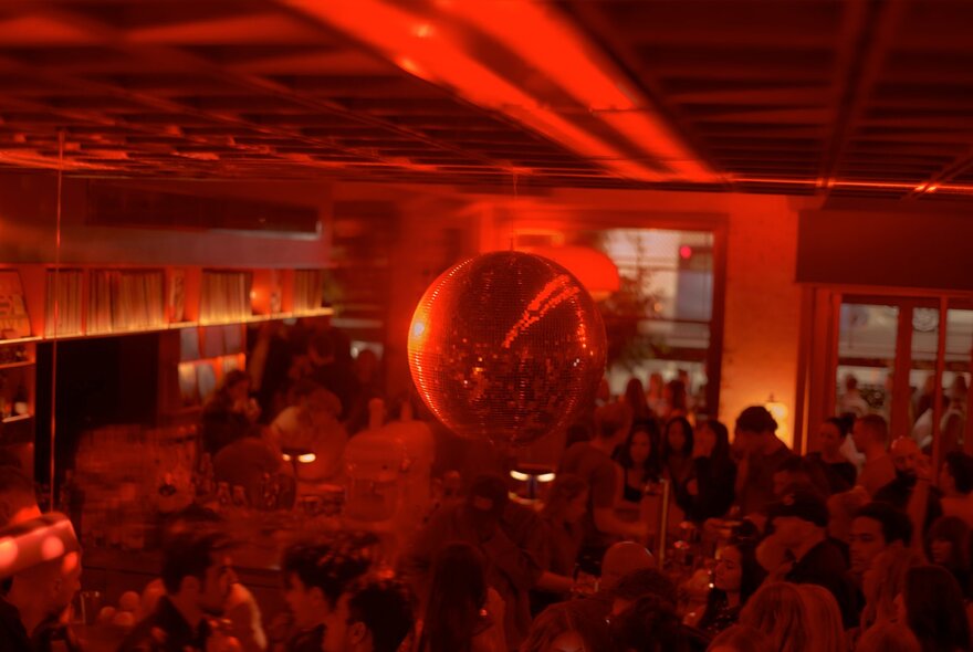 Looking down at the bar counter at LB's Record Bar, with people drinking and socialising, under a large mirror ball, with warm and atmospheric reddish lighting.