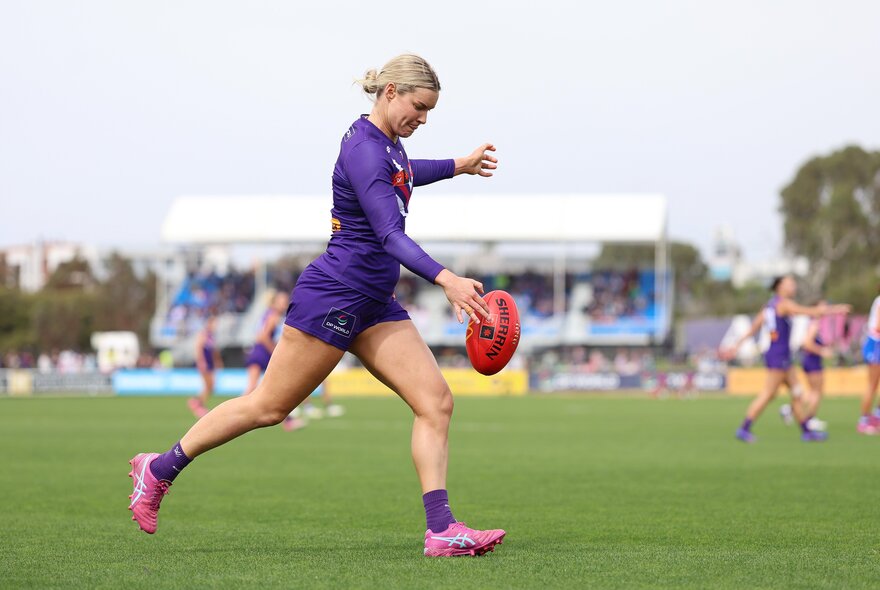 A Fremantle AFLW player running on the field with the football in her hands about to kick it.