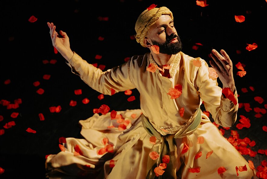 Dancer Aakash Odedra wearing sand coloured robes and a head scarf, holding a dance pose on a stage, surrounded by falling red rose petals.