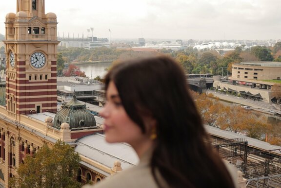 Blurred head of a person in front of Flinders St Station clock tower, Melbourne railyards and stadiums in the background.