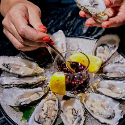 Hand hovering over a plate of shucked oysters, selecting one with a small fork and dippin it into a bowl of sauce.