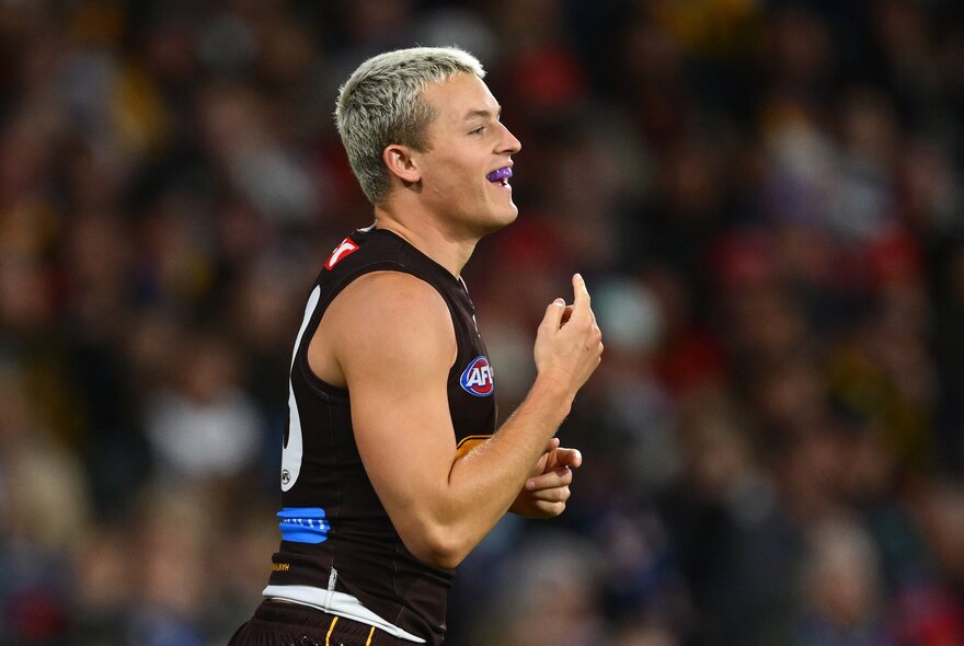 An AFL football player smiling during a match, a blurred crowd of stadium fans in the background.