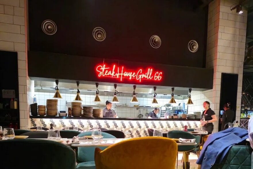 Looking over tables with coloured velvet chairs towards the kitchen at Steakhouse Grill 66, where an opening is half filled with plates.