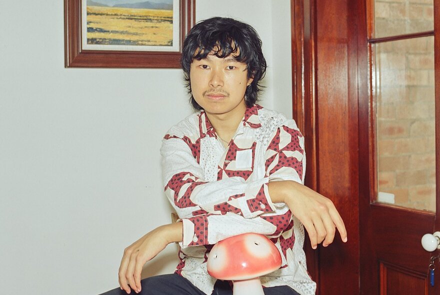 A man with arms crossed as he leans on a replica red toadstool, in a room with an old wooden door with glass inlay. 