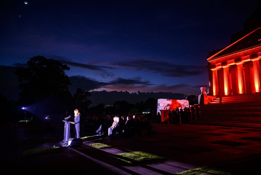 A person standing at a podium in front of the Shrine for the ANZAC Day Dawn Service.