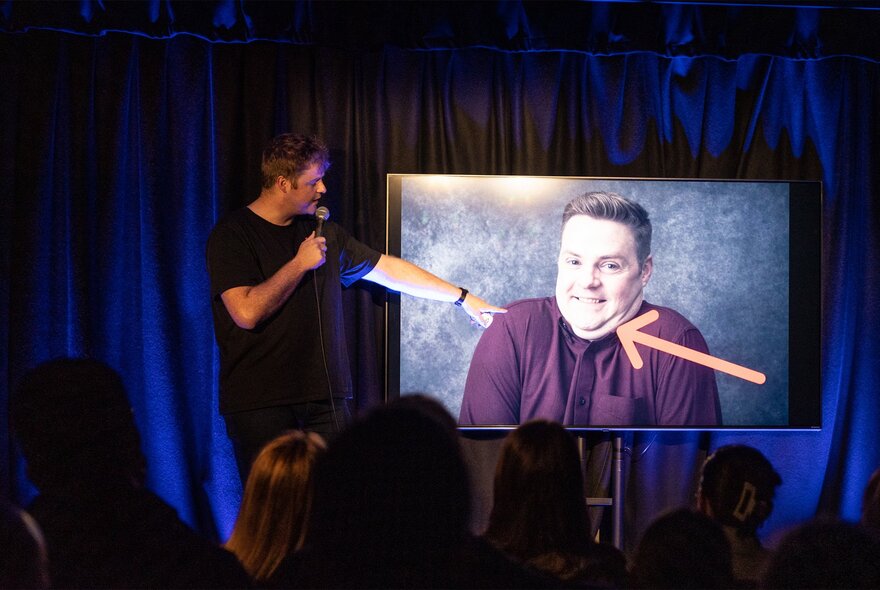 A performer on stage, talking into a microphone he holds, while pointing to an image of himself projected on a screen behind him.