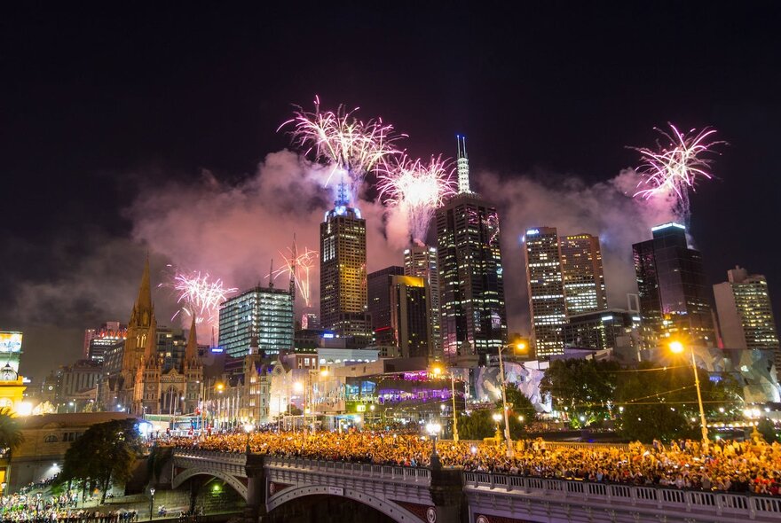 Fireworks above Melbourne's city buildings with crowded Princes Bridge vantage point.