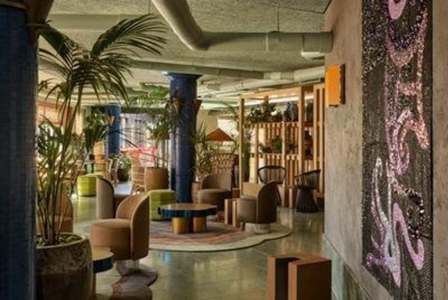 A lush bar and lounge area with modern style seating, many large potted plants, a round coffee table, concrete pillars, and abstract art on the wall.