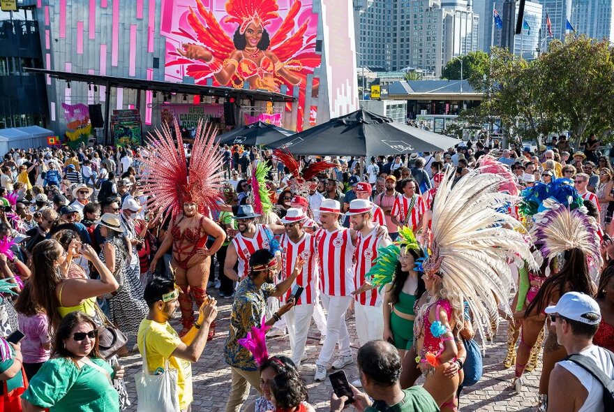 Dancers wearing elaborate Carnival costumes performing in the centre of a crowd at Fed Square, at an outdoor daytime festival.