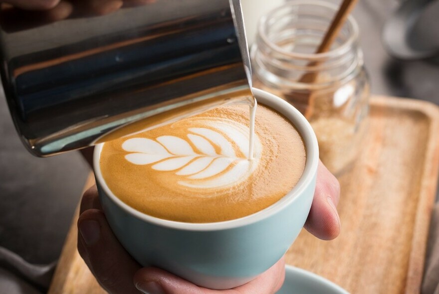 Barista's hand holding a cup of coffee, pouring in milk in a pattern.