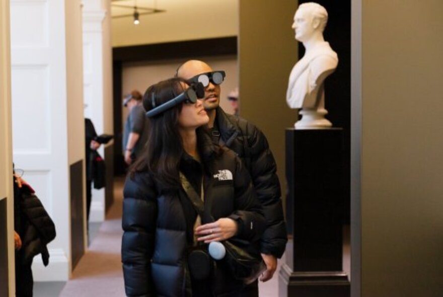 People at an exhibition wearing VR headgear, a white bust on a plinth in the background. 