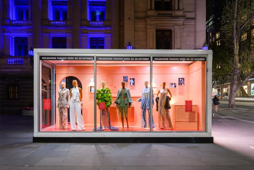 A Melbourne Fashion Week outdoor fashion capsule installation of fashion mannequins in a large glass rectangular case, displayed in a public space.