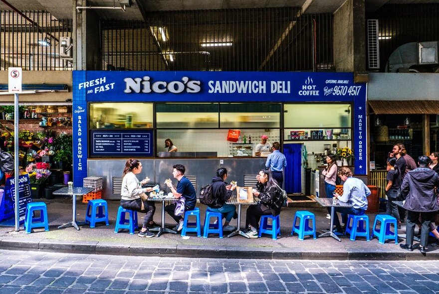 People dining on blue stools outside a bright blue sandwich deli.