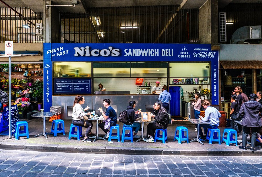 People dining on blue stools outside a bright blue sandwich deli.
